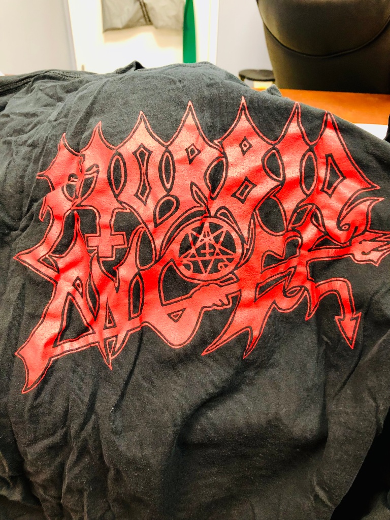 A photograph of a wrinkled and faded black T-shirt with the name of the heavy metal band Morbid Angel spelled out in red, angular, pointed block letters across the middle of the shirt. This font morphs together in a graffiti style, and several letters form symbols associated with this genre of music –on the bottom right leg of the M, there is an upside down cross; in the middle section of the letter G, there is a pentagram; at the bottom of the text a pitchfork, held by a hand extending from the base of the letter G underlines the word "angel"; and both the D and L at the end of each word has a devil's tail.